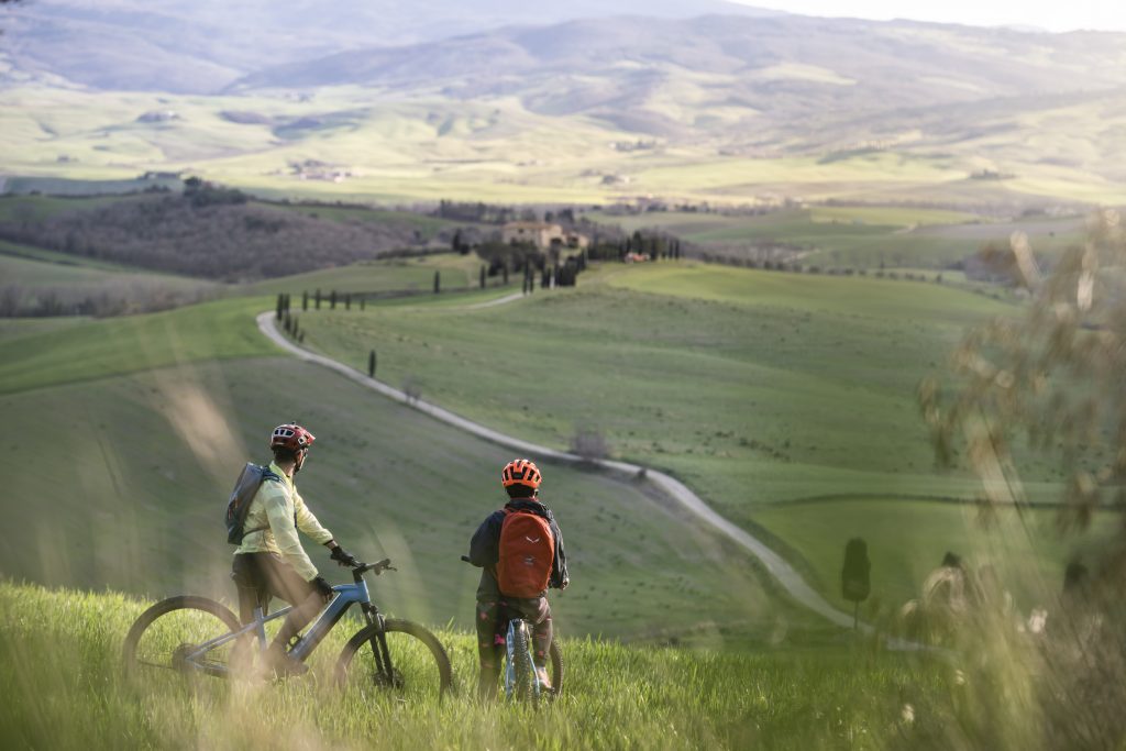 Due cicloturisti in primo piano con sull osfondo campagna toscana e cipressi